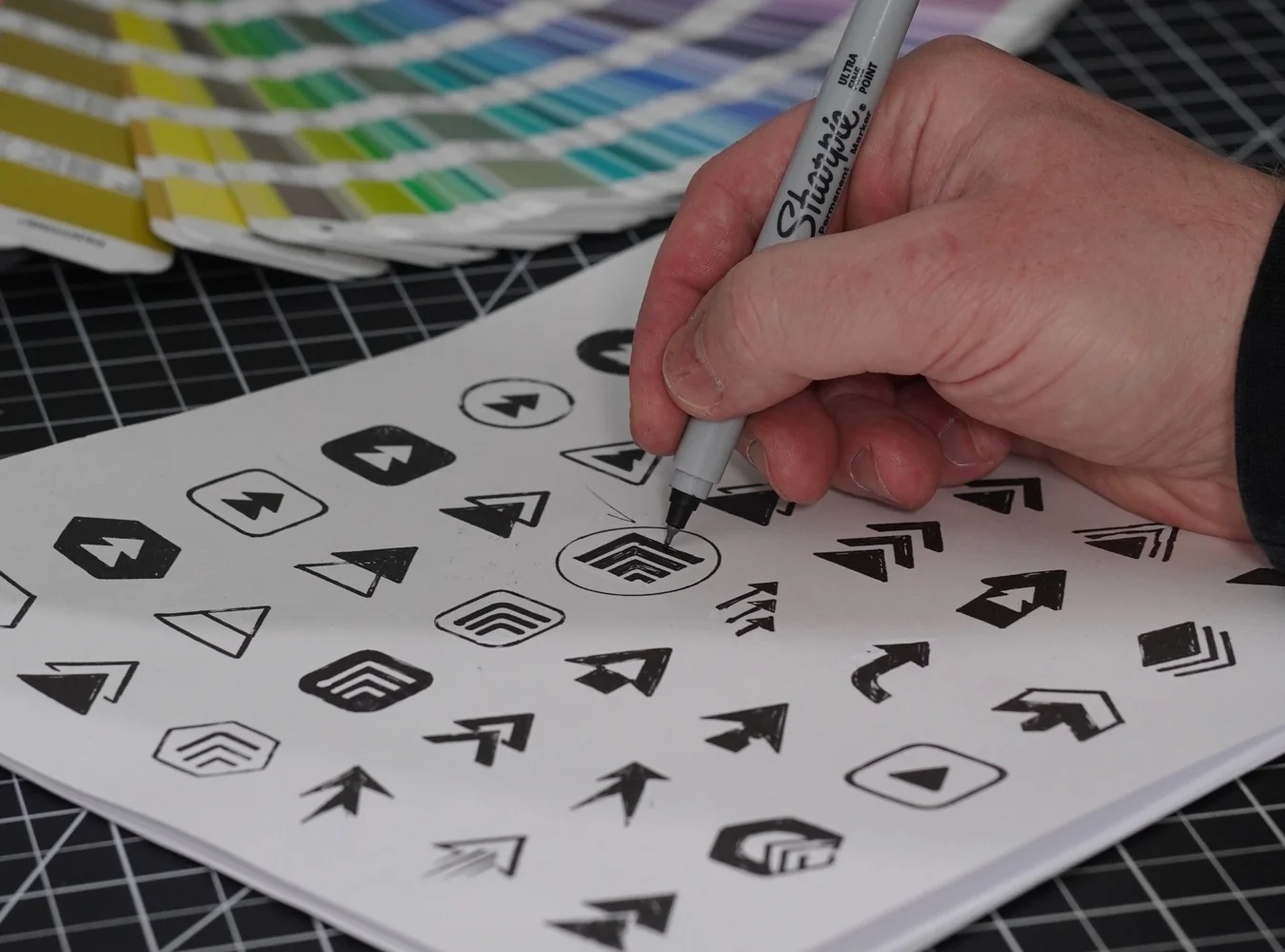 Close-up of a hand refining arrow-shaped logo sketches with a Sharpie beside color swatches